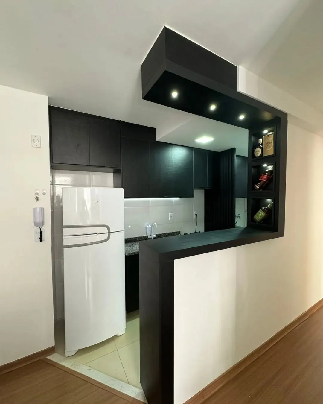 Cozinha compacta com armários pretos, bancada com nichos iluminados para garrafas e geladeira branca