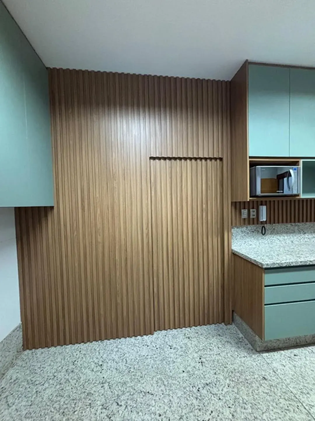 Painel ripado de madeira com porta camuflada, ao lado de armários verde-sálvia