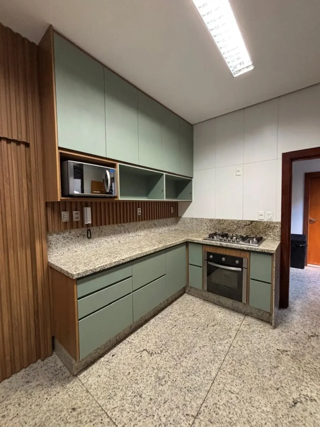 Cozinha em L verde-sálvia com bancada de granito, forno embutido e painel ripado ao lado